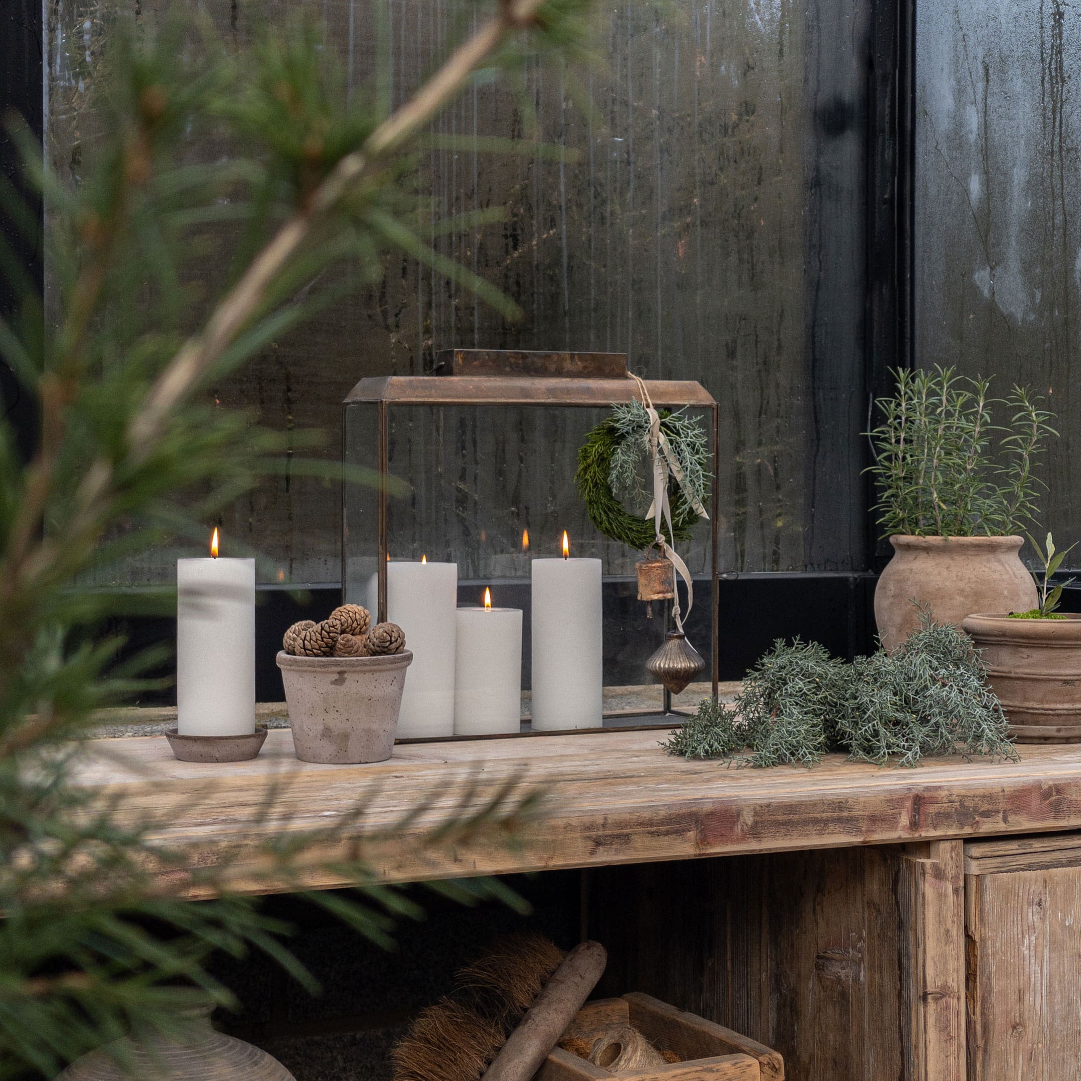 Rustikaler Holztisch mit weißen Kerzen und Tontöpfen mit grünen Pflanzen vor beschlagener Scheibe