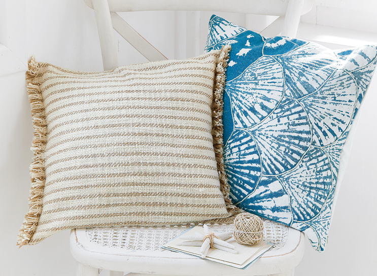 Zwei dekorative Kissen auf weißem Stuhl, eines beige gestreift mit Fransen, das andere blau mit Muschelmotiv