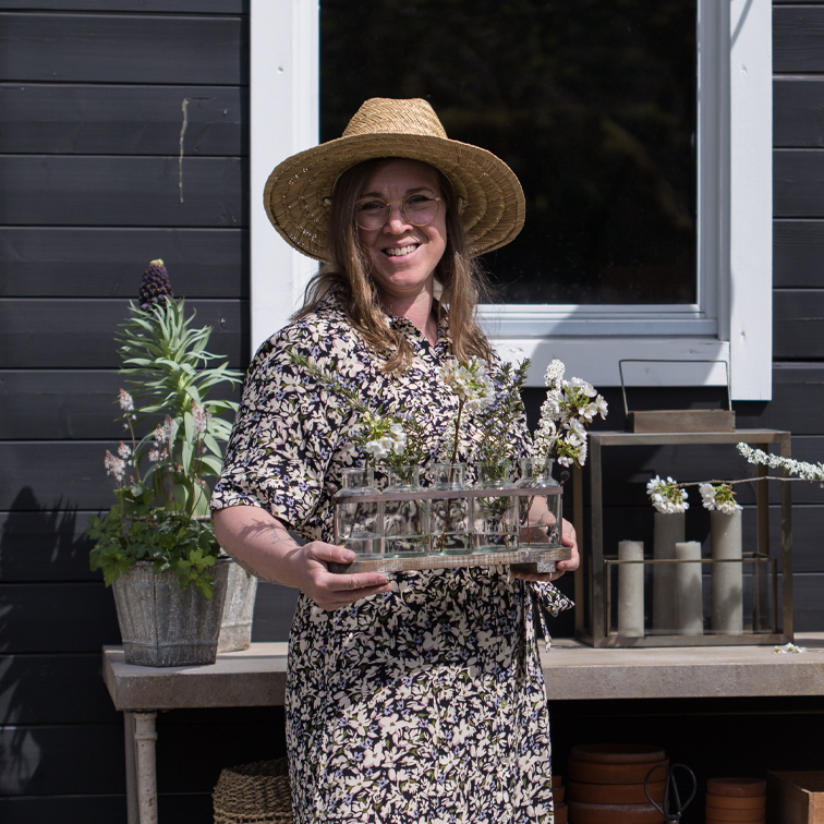 Frau mit Strohhut hält Dekoration aus Glas-Reagenzgläsern mit Blumen vor schwarzer Wand