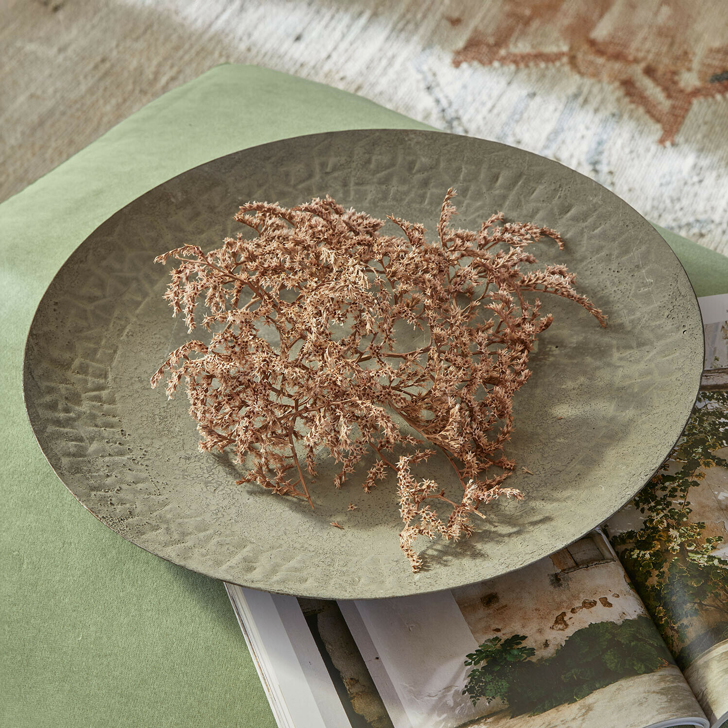 Große grüne Metall-Dekoschale mit Struktur und getrockneten Blumen auf grünem Stoff und Magazin