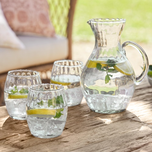 Glaskrug und drei Gläser mit Wasser, Zitrone und Minze auf Holztisch im Freien