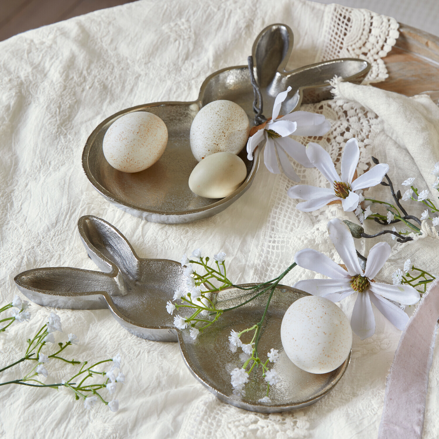 Metalltablett in Hasenform mit weißen Eiern und Blumen auf hellem Tuch, österliche Dekoration