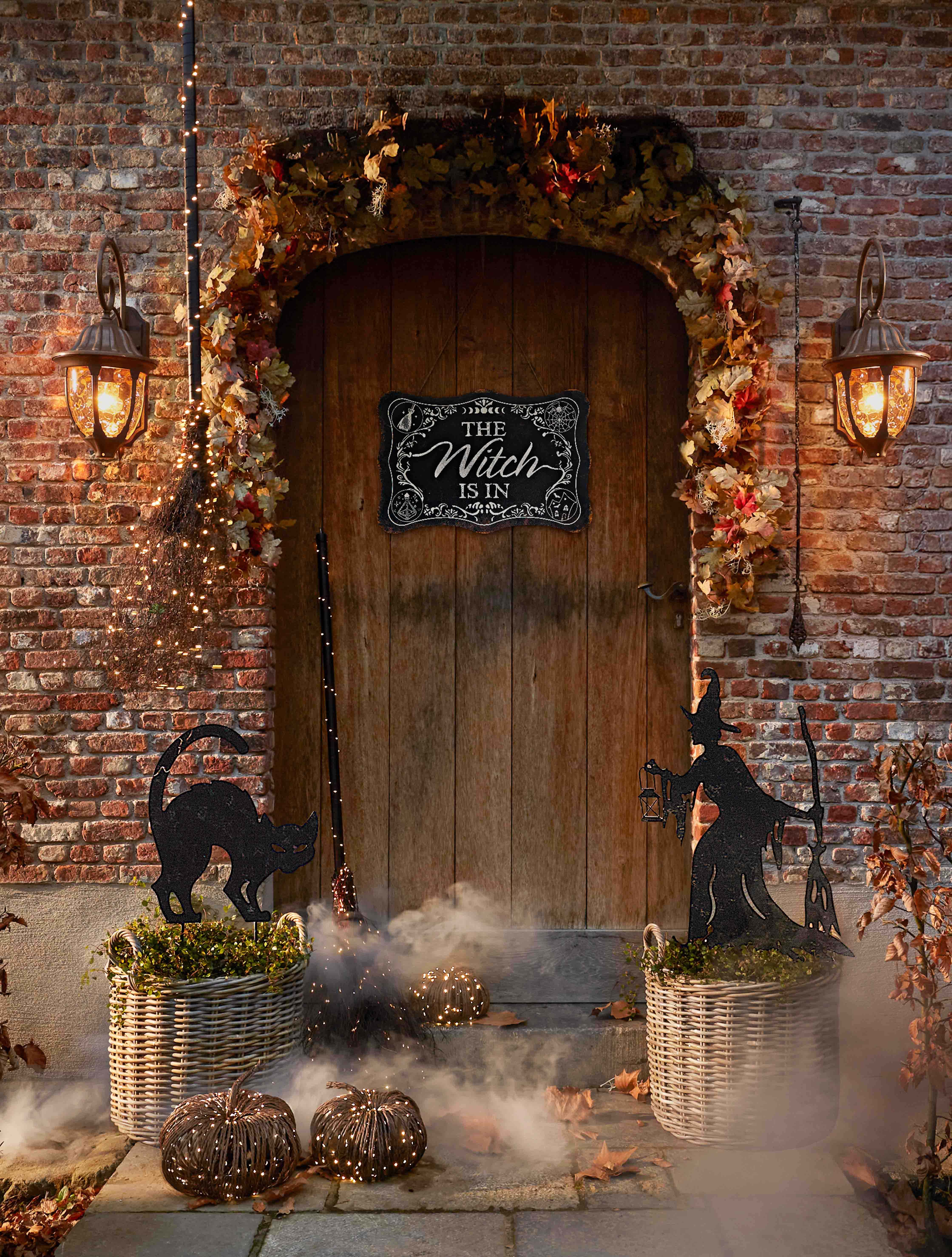 Halloween-Dekoration mit Hexe, schwarzer Katze und Laternen vor Holztür und Ziegelwand