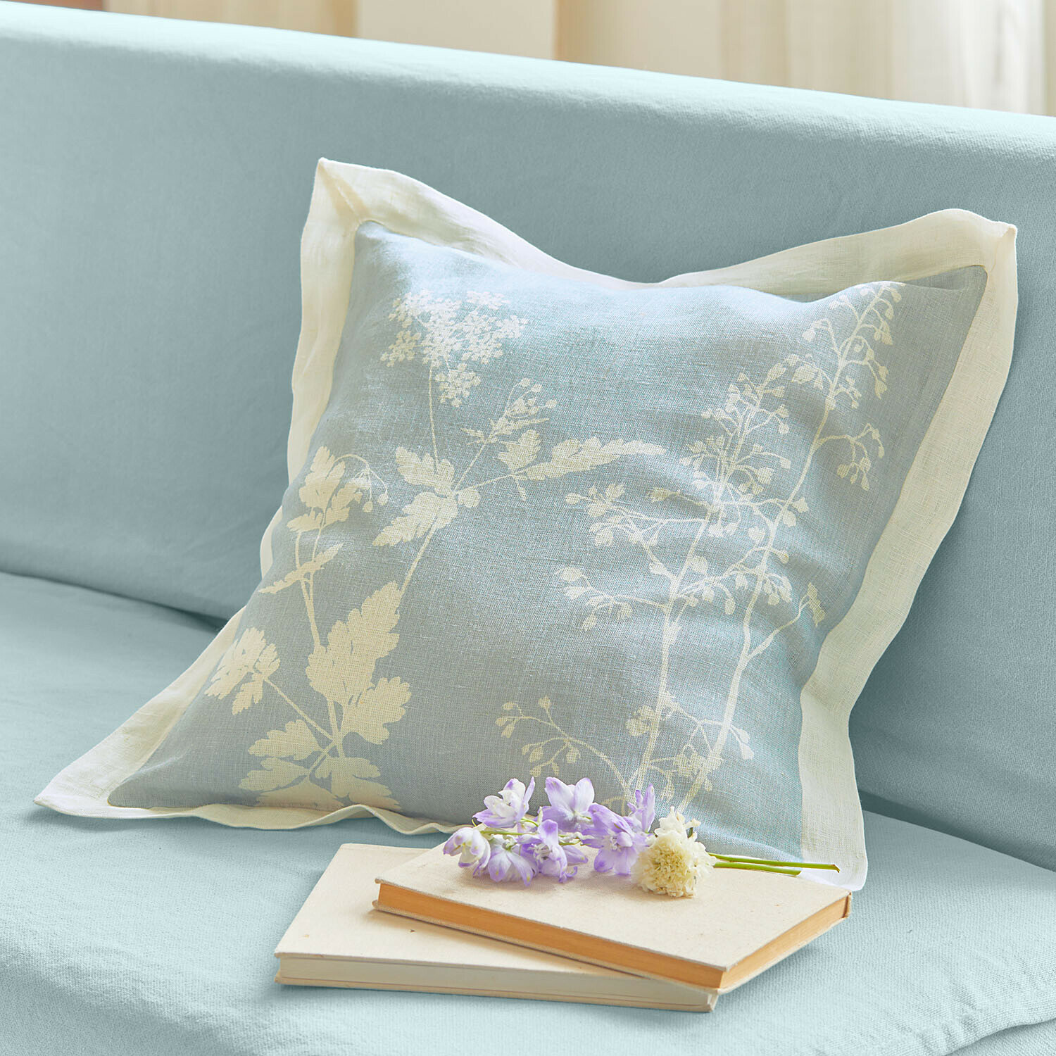 Hellblaues Leinenkissen mit weißem Blumenmuster auf Sofa, daneben Bücher und Blumen