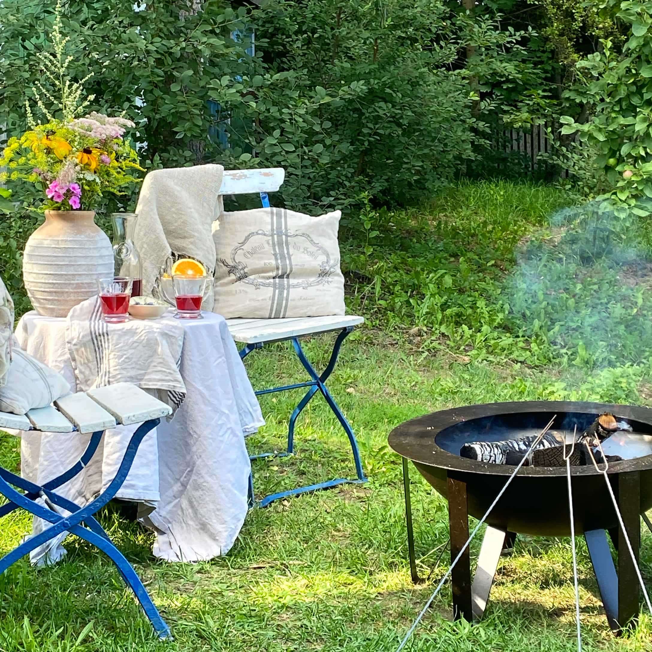 Garten mit blauen Metallstühlen, weiß gedecktem Tisch und schwarzem Feuerkorb mit rauchendem Holz