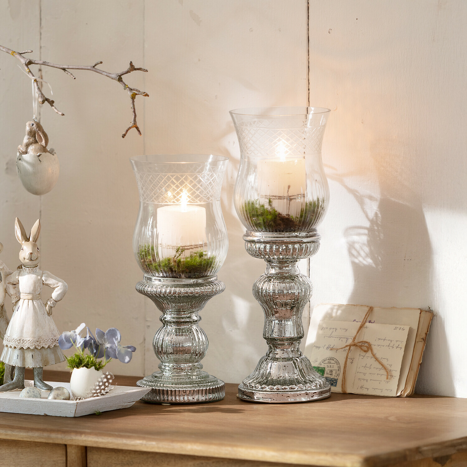 Zwei Glas-Kerzenhalter mit weißen Kerzen und grünem Moos auf Holztisch, Frühlingsdekoration