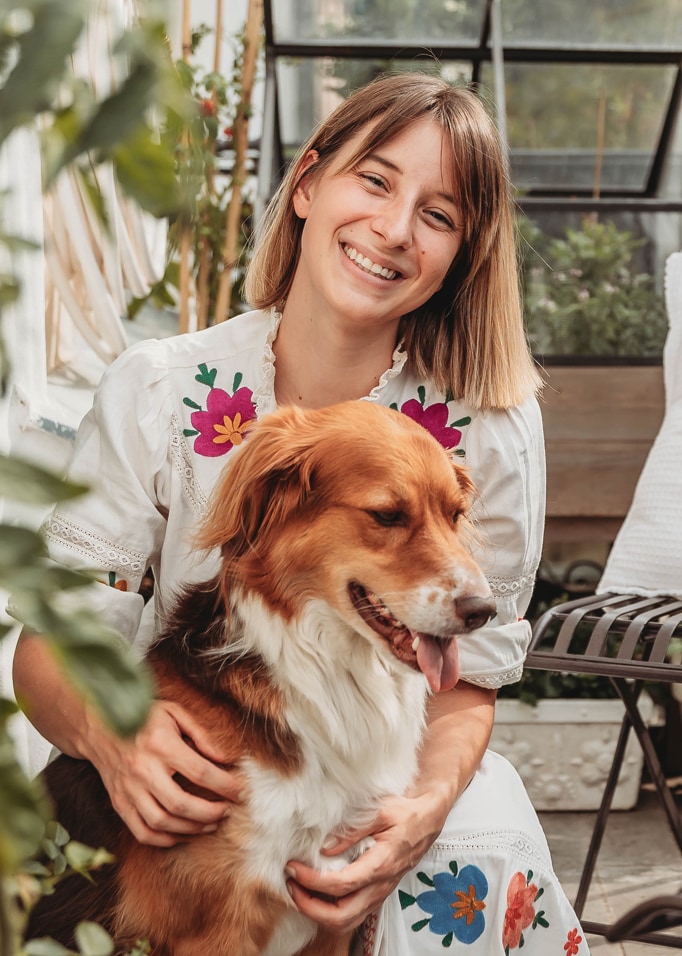 Frau in weißem Kleid mit bunten Stickereien hält braun-weißen Hund in hellem Raum mit Pflanzen
