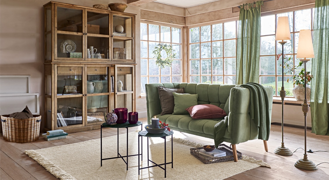 Gemütliches Wohnzimmer mit grünem Stoffsofa, Holzvitrine und natürlichem Teppich auf Holzboden