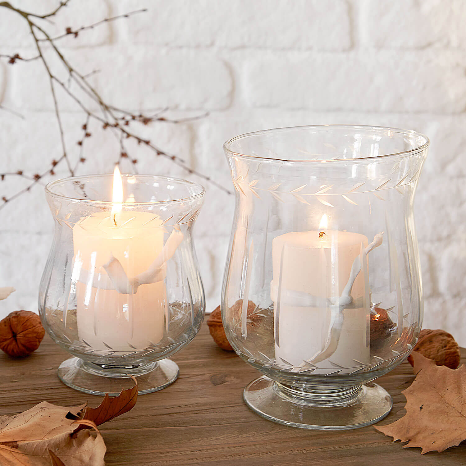 Zwei durchsichtige Glaswindlichter mit weißen Kerzen auf Holztisch mit Herbstblättern dekoriert