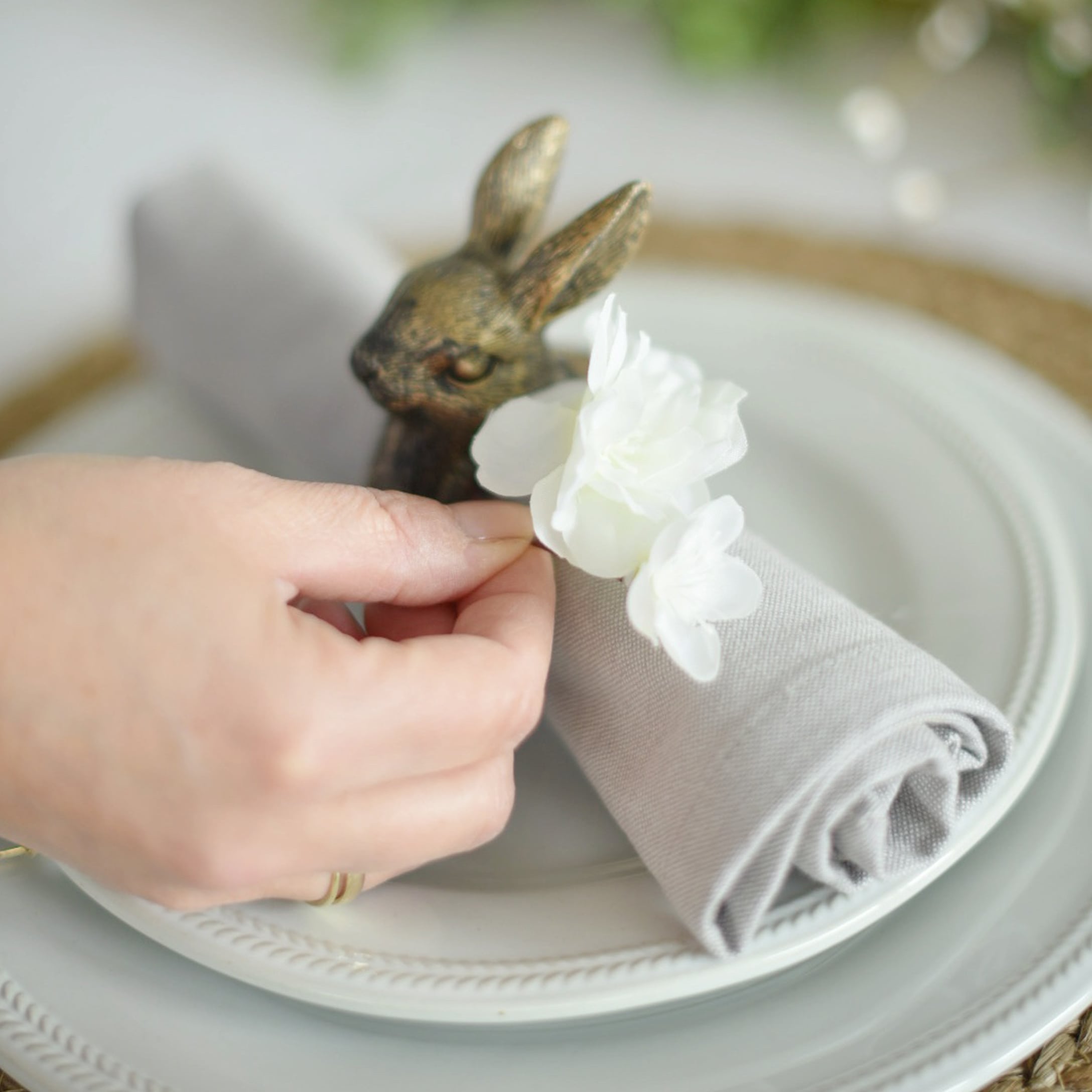 Grauer Leinen-Serviettenring mit weißer Blume und Messing-Hasendekor auf weißem Teller