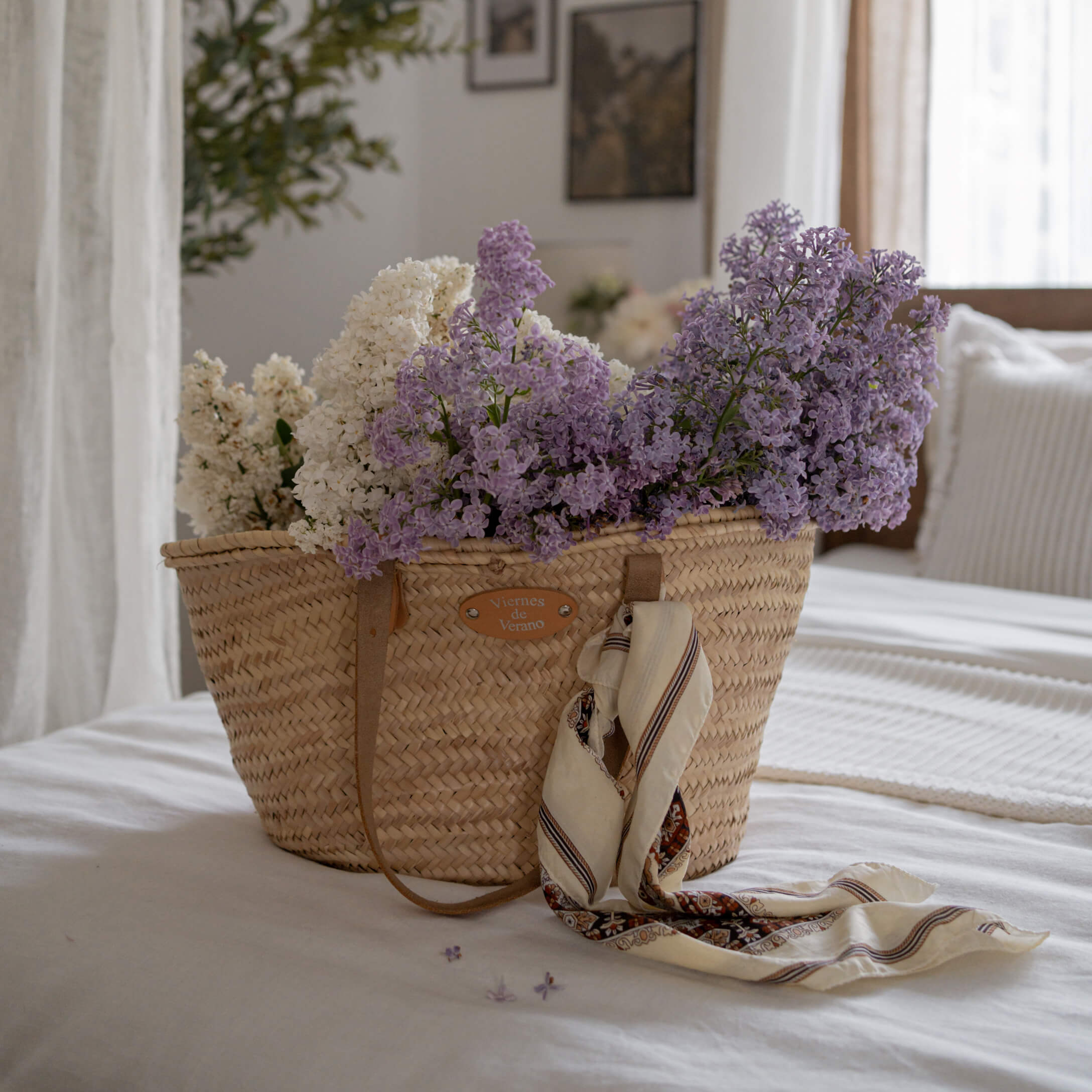 Geflochtener Korb mit weißen und violetten Blumen und Tuch auf weißem Bett in hellem Zimmer