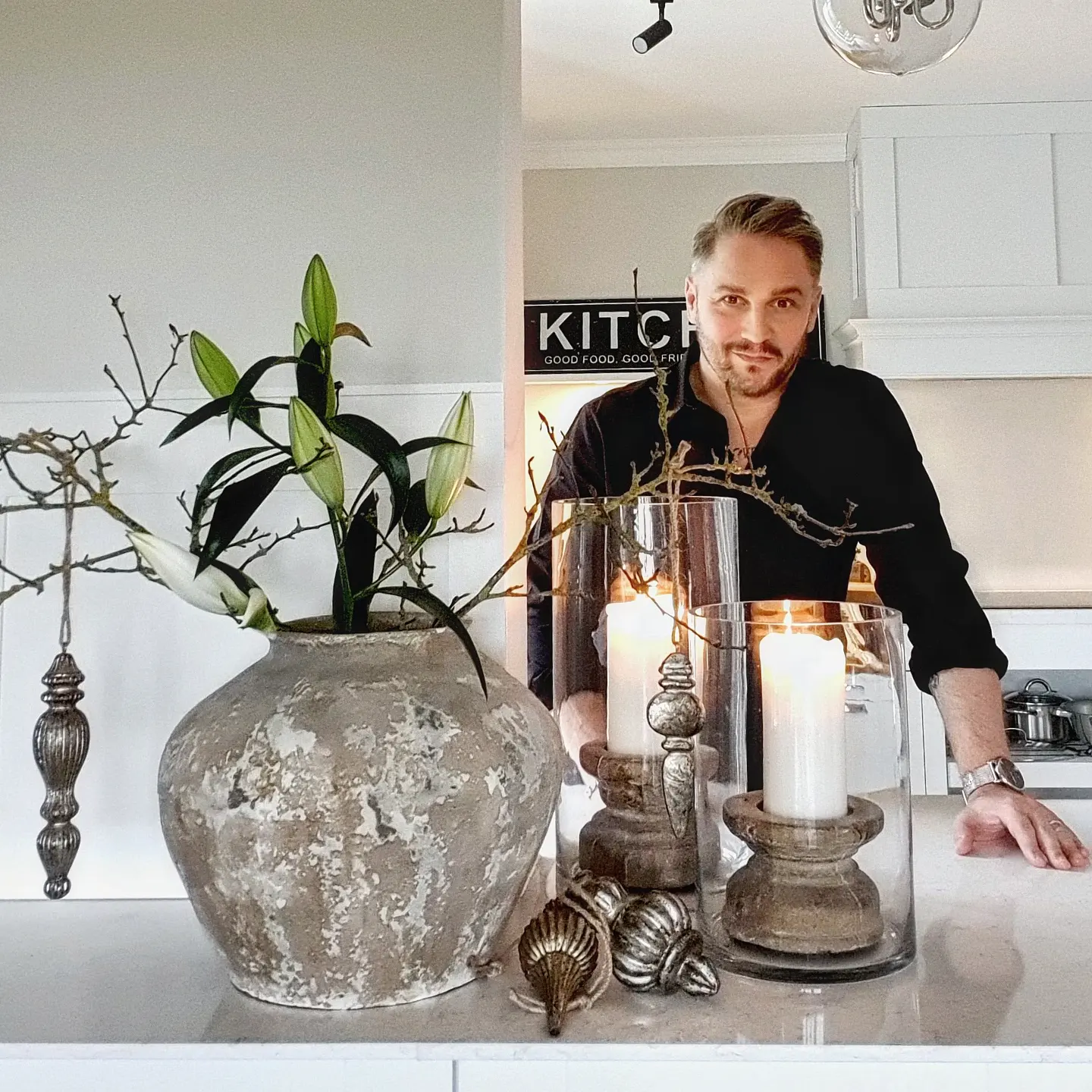 Große graue Keramikvase mit grünen Blättern und brennenden Kerzen in Glaswindlichtern auf Küchenarbeitsplatte