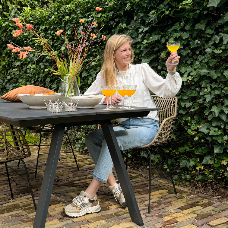 Frau in weißer Bluse und Jeans sitzt draußen an schwarzem Tisch, hält Glas mit orangem Getränk, Vase mit Blumen daneben