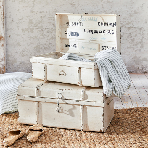 Weiße vintage Holzkisten mit Gebrauchsspuren, Textilien und Hausschuhen auf Juteteppich