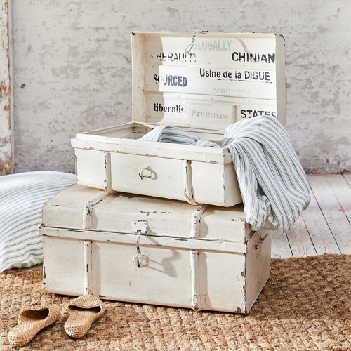 Weiße vintage Holzkisten mit Gebrauchsspuren, Textilien und Hausschuhen auf Juteteppich