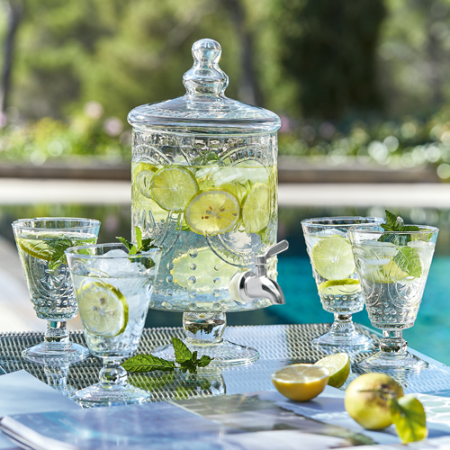 Glasgetränkespender mit Zitronenscheiben und Minze, vier Gläser auf Gartentisch am Pool