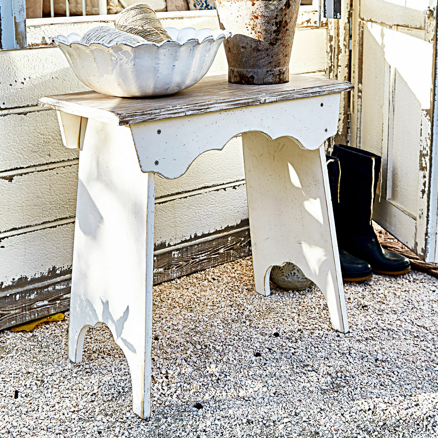 Hocker Fria Weißer Holztisch mit Shabby-Chic-Oberfläche auf Kiesboden, dekorative Schale und schwarze Stiefel
