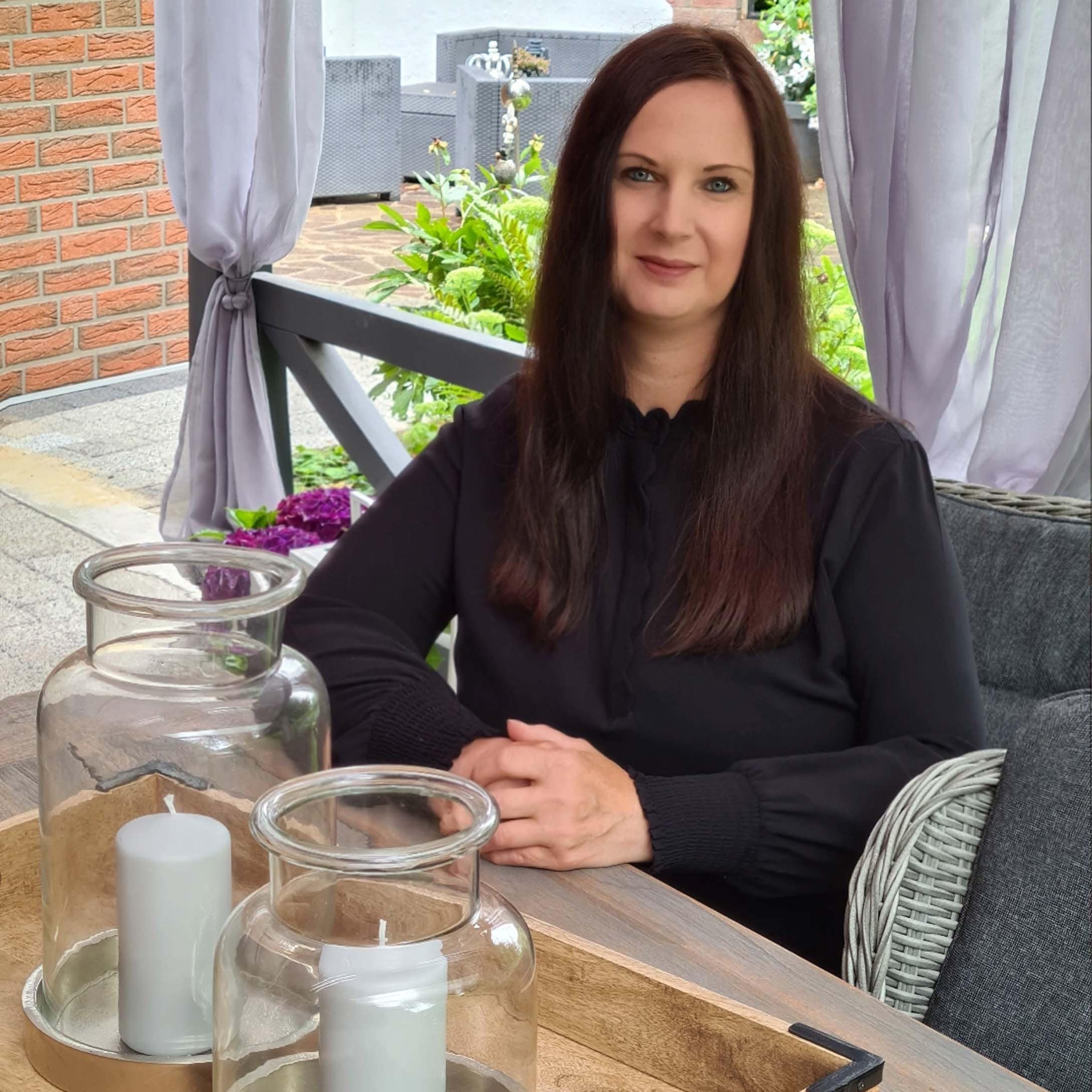 Frau in schwarzer Bluse sitzt an Holztisch mit dekorativen Kerzen in Glaslaternen auf Terrasse
