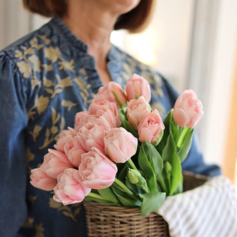 Strauß rosa Tulpen in einem Weidenkorb gehalten von Frau in blauem Blusenhemd mit Blumenmuster
