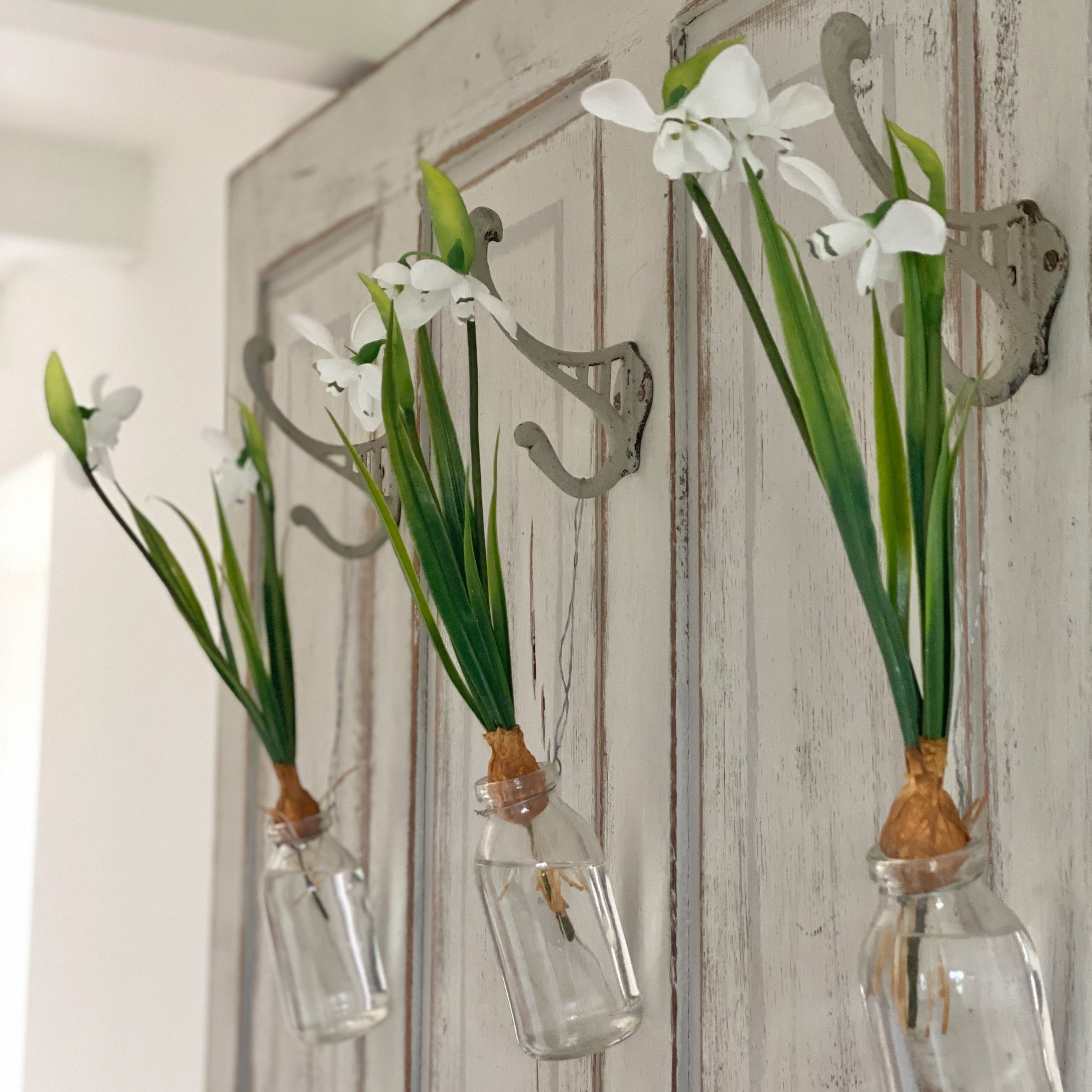 Drei weiße Blumen in Glasflaschen an Metallhaken an einer Holztür hängend