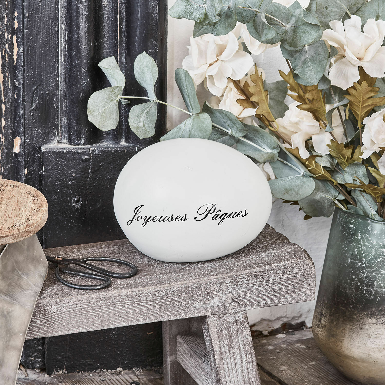 Deko-Ei JOYEUSES PAQUES Weißes Dekoei mit Schriftzug Joyeuses Pâques auf Holzbank mit Blumen und Schere