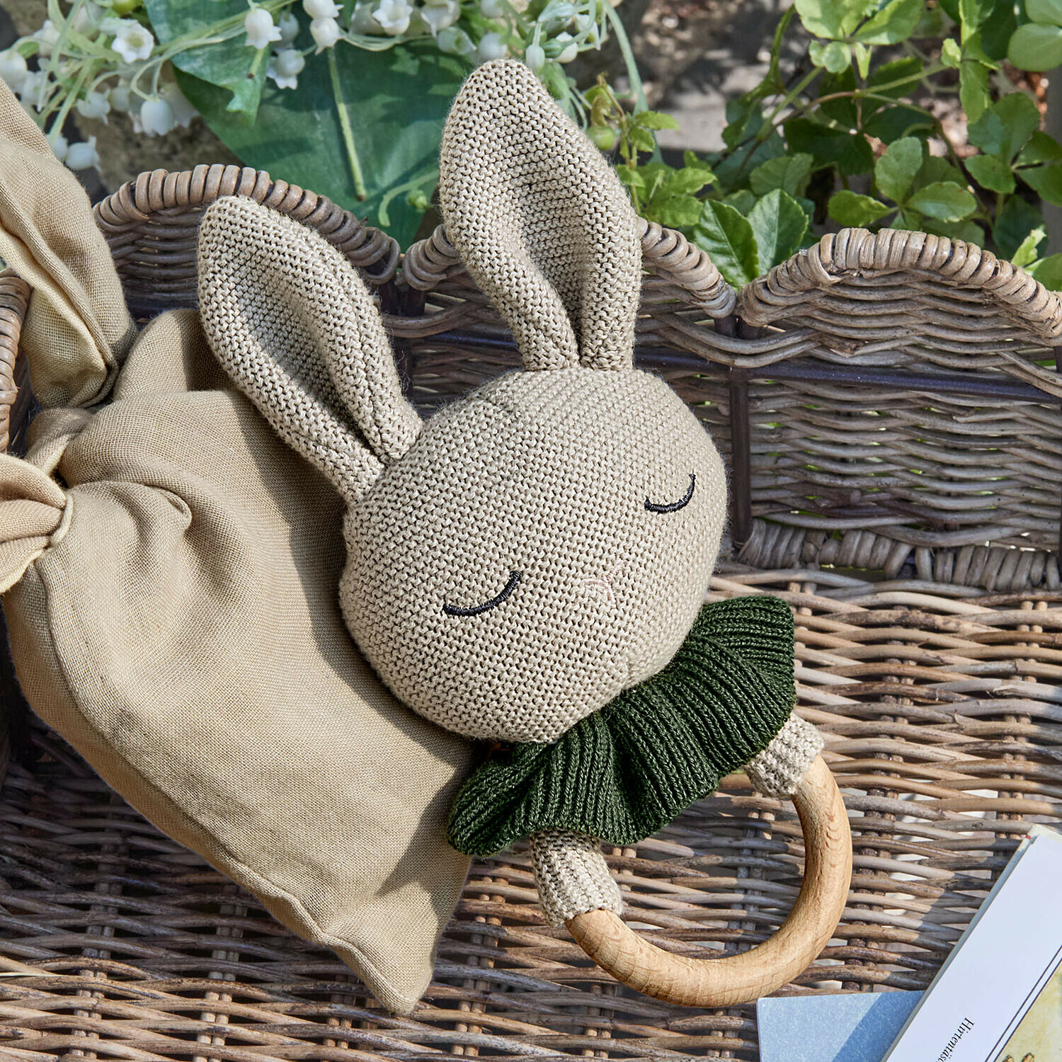 Spielzeugring Cuddlecloud Beige gestrickte Hasenrassel mit grünem Kragen und Holzring auf Korbhintergrund
