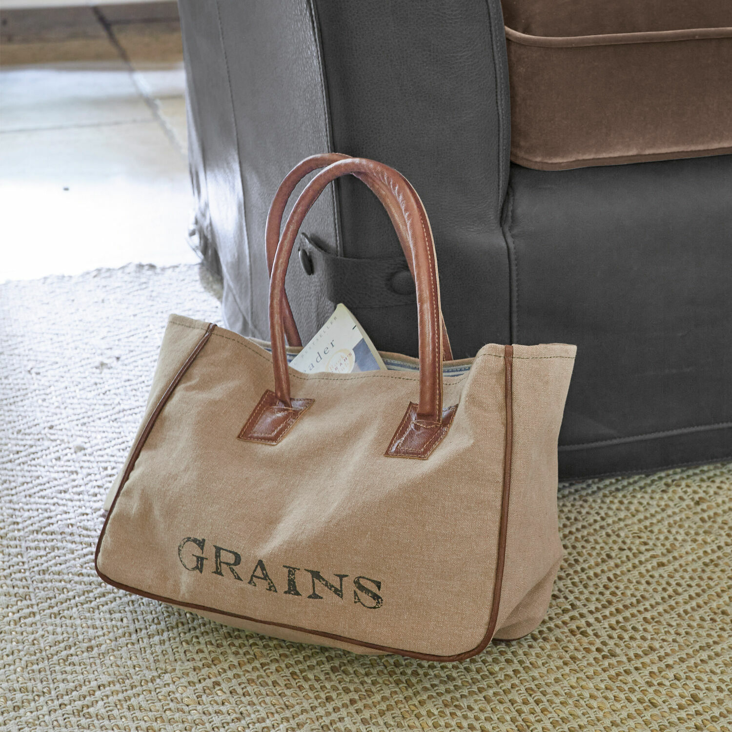 Tasche Grains Beige Stofftasche mit braunen Ledergriffen und Schriftzug GRAINS auf Teppich neben Sofa