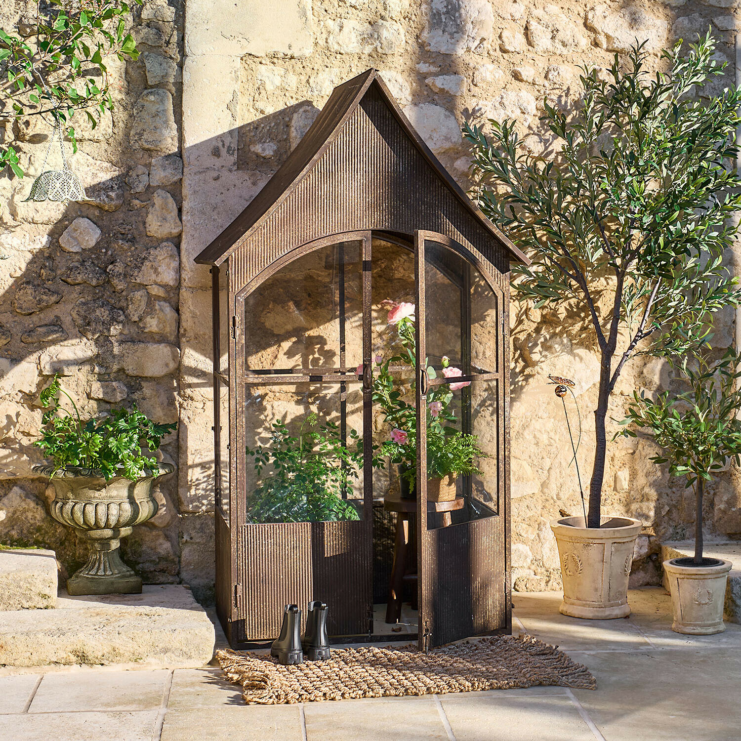 Vintage Metall-Gewächshaus in Braun mit Pflanzen und Töpfen vor einer sonnigen Steinmauer im Garten