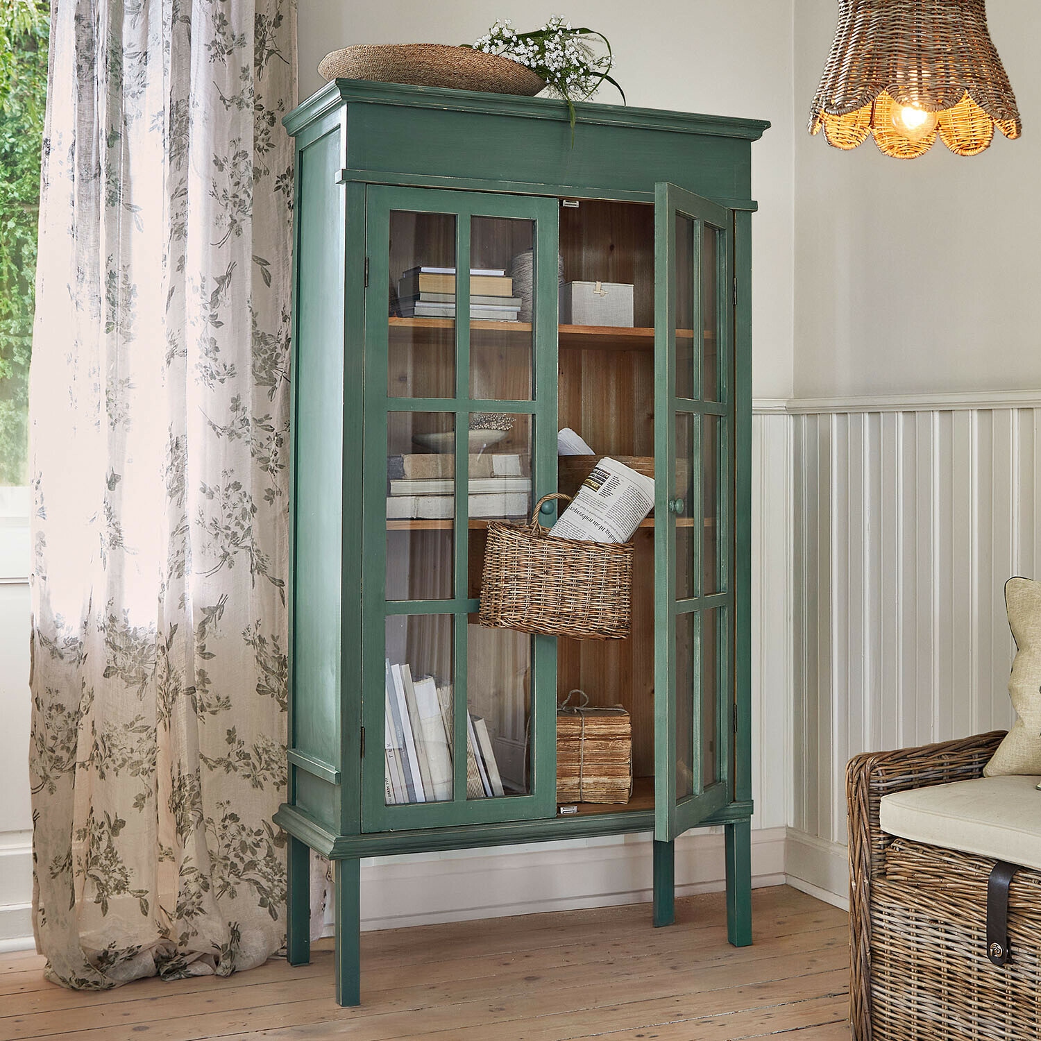 Grüner Holzvitrinenschrank mit Glastüren und Korb aus Weide im hellen Wohnzimmer mit natürlichem Licht