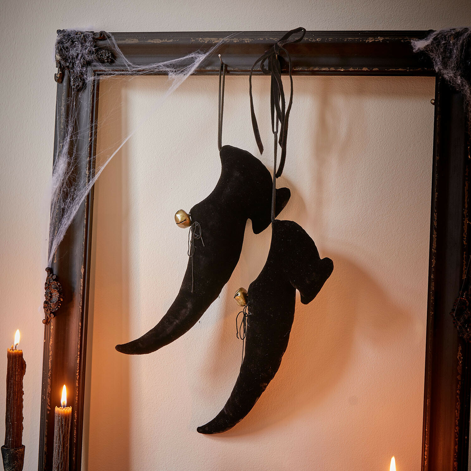 Schwarze Samt-Hexenschuhe mit Glöckchen, hängend an einem antiken Rahmen mit Spinnweben und Kerzen