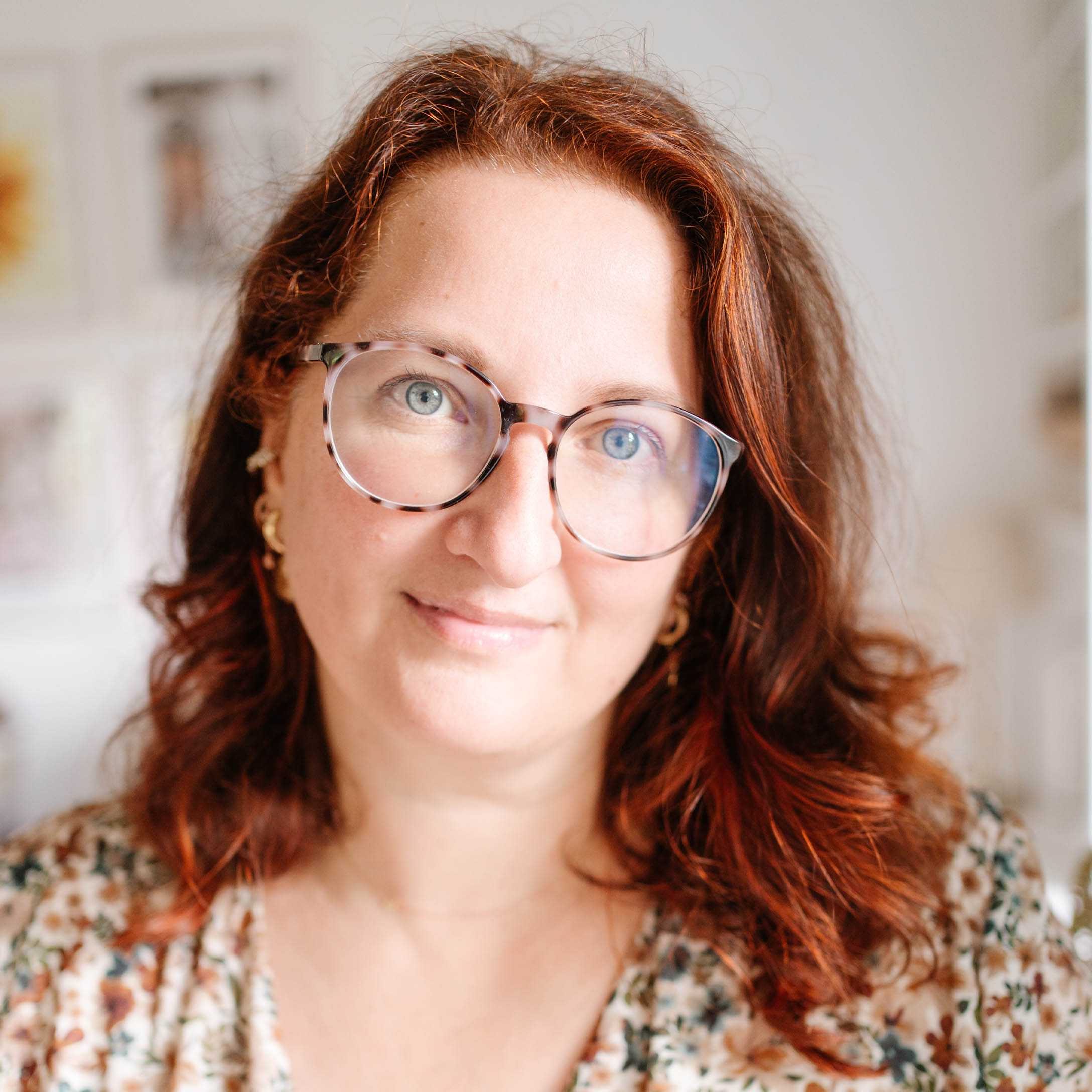 Porträt einer Frau mit roten Haaren und Brille in geblümter Bluse vor hellem Hintergrund