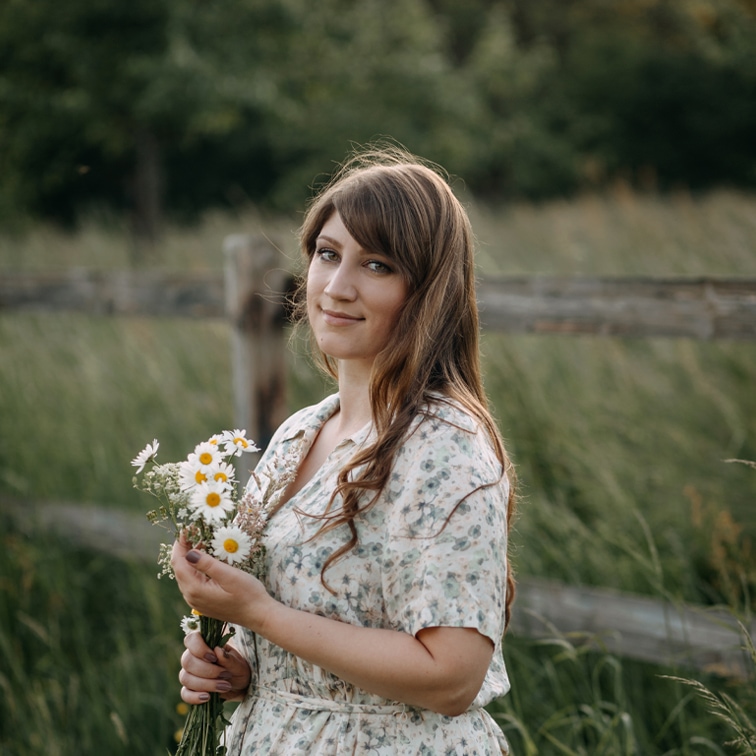 Frau im geblümten Kleid hält einen Strauß Gänseblümchen vor grünem Feld und Holzzaun
