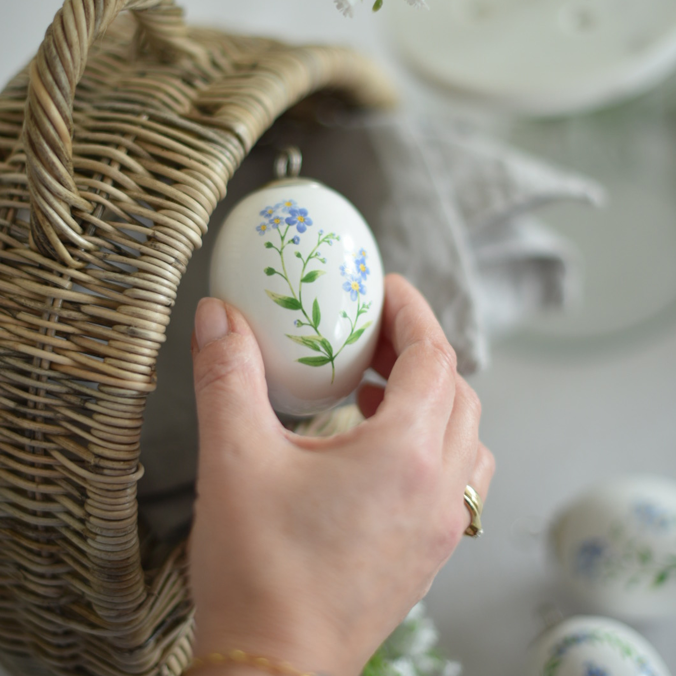 Hand hält weiße Keramik-Ostereier-Dekoration mit blauen Blumen neben einem Weidenkorb