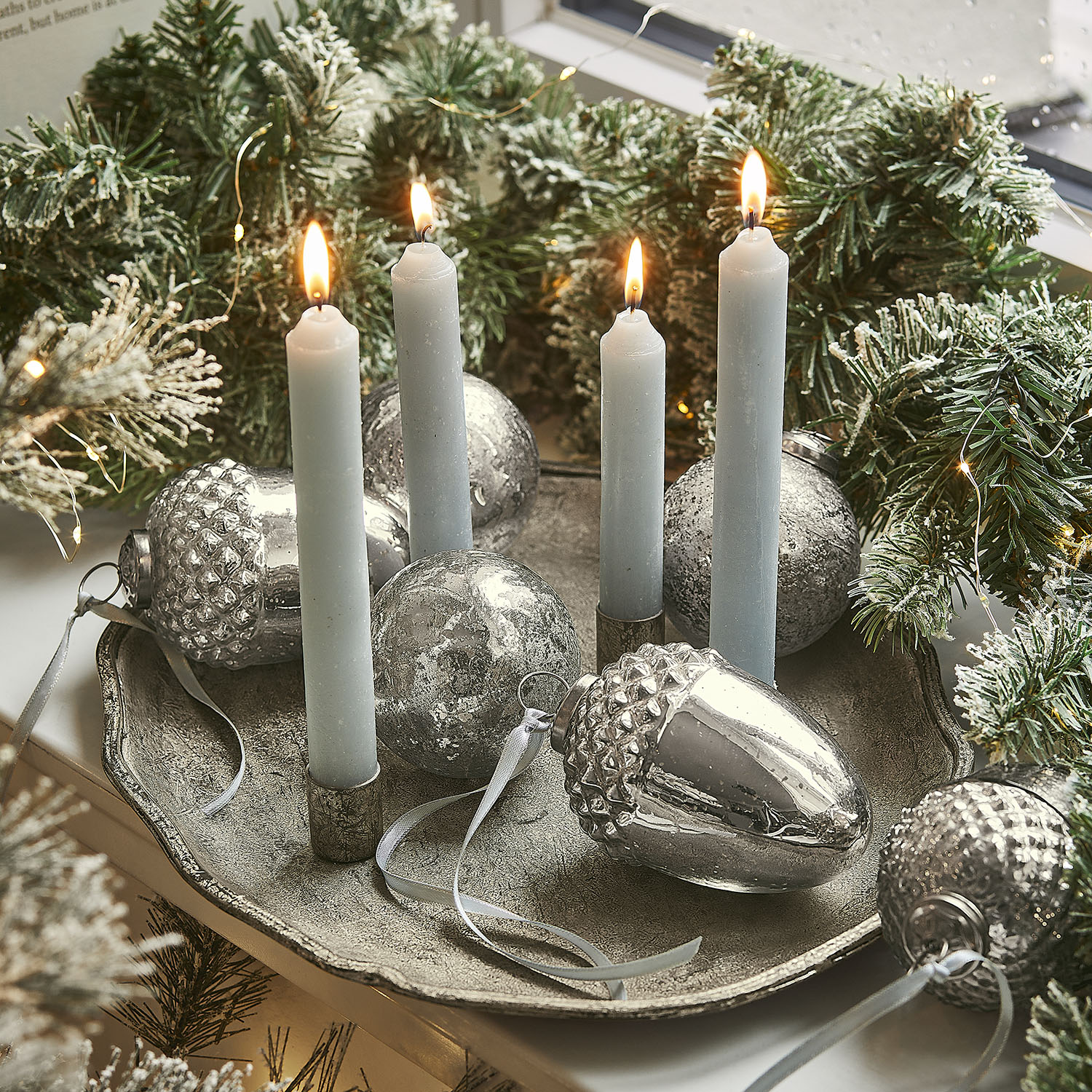 Kerzenhalter mit vier blauen Kerzen und silbernen Dekorationen auf Metalltablett mit grünen Zweigen