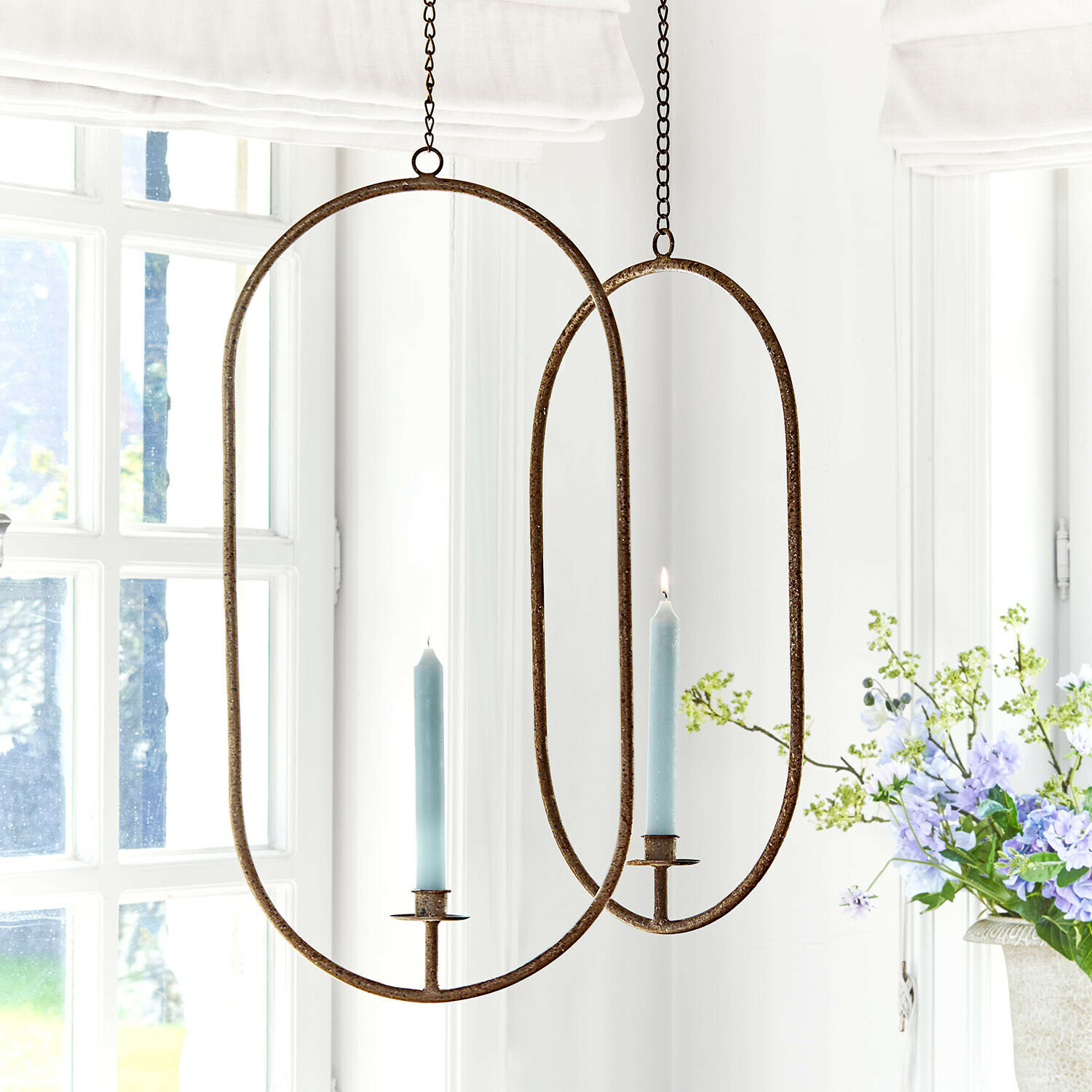 Ovaler Metall-Hängeleuchter mit blauer Kerze in hellem Raum mit Blumenarrangement