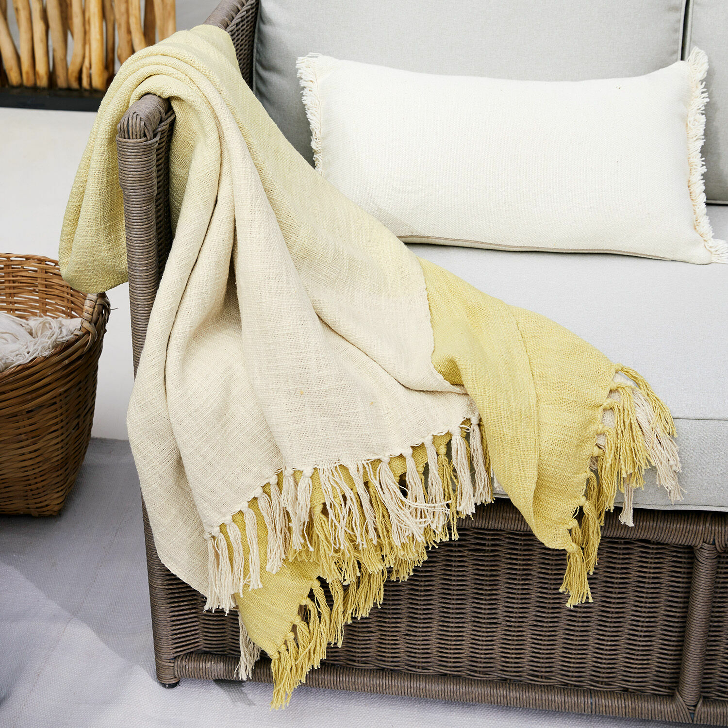Plaid Aurice Zweifarbige Baumwolldecke in Creme und Gelb mit Fransen auf grauem Rattansofa